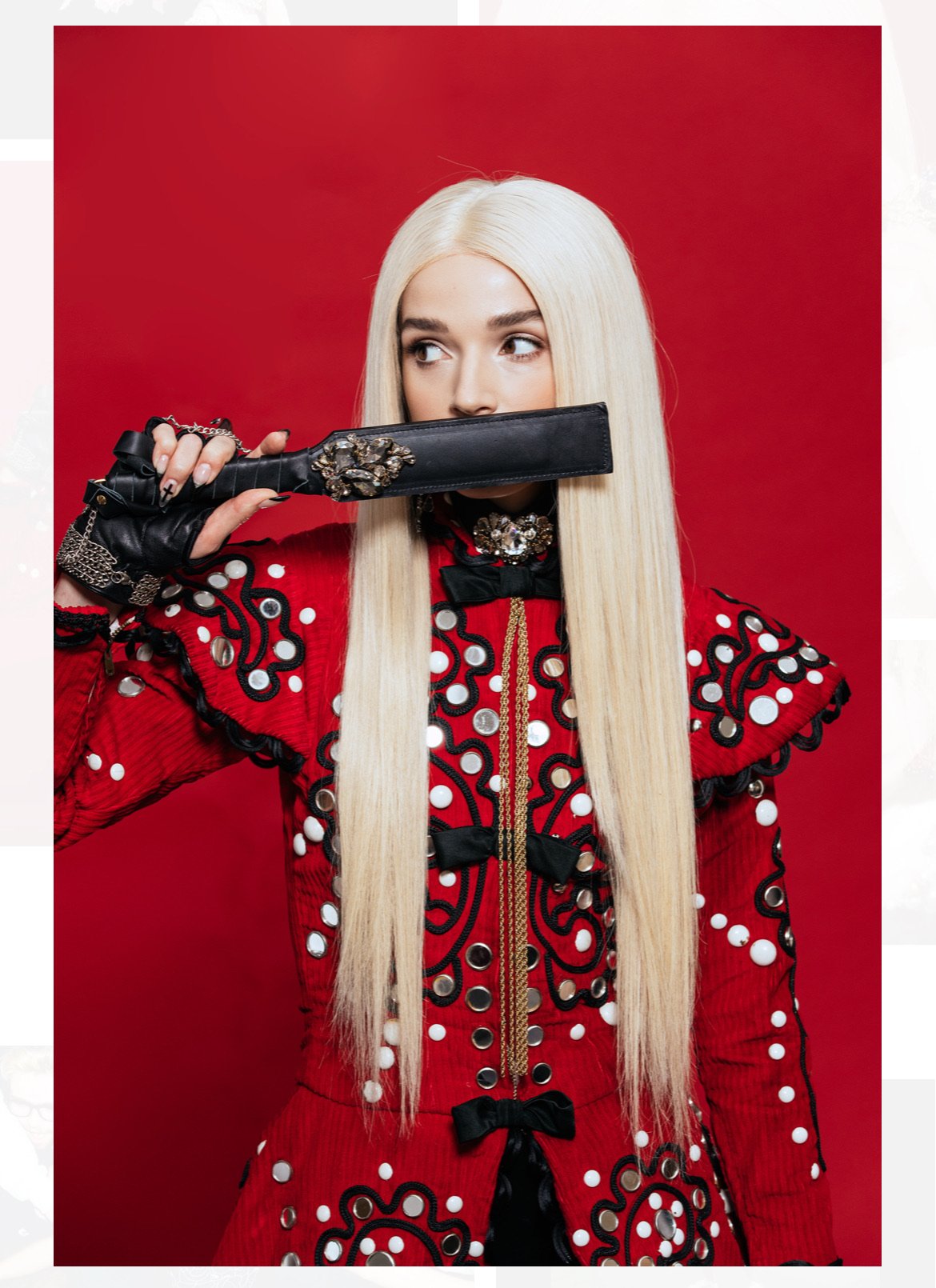 Portrait of adult female with long blonde hair, wearing a red jacket with white accents, holding a black object against a red background.