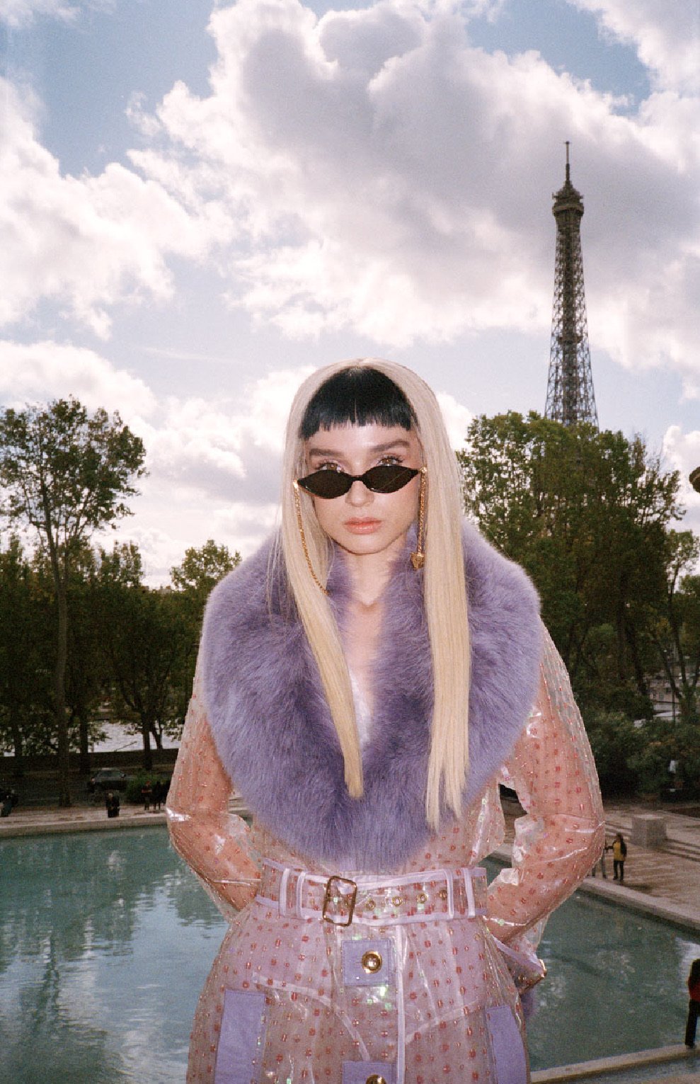 Adult female with sunglasses and blonde hair wearing lilac fur collar coat in front of the Eiffel Tower in Paris on a sunny day.