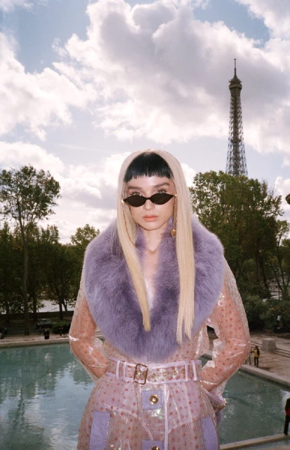 Adult female with sunglasses and blonde hair wearing lilac fur collar coat in front of the Eiffel Tower in Paris on a sunny day.