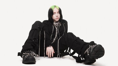 Fashionable adult female, Billie Eilish, sits on a white background wearing a black jacket, pants, jewelry, and spiked platform shoes.