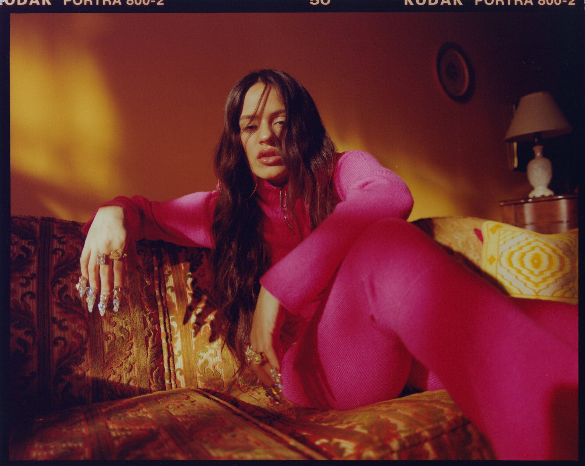 Stylish woman in a pink jumpsuit poses on an ornate couch in an indoor setting with warm lighting and film grain effect visible.