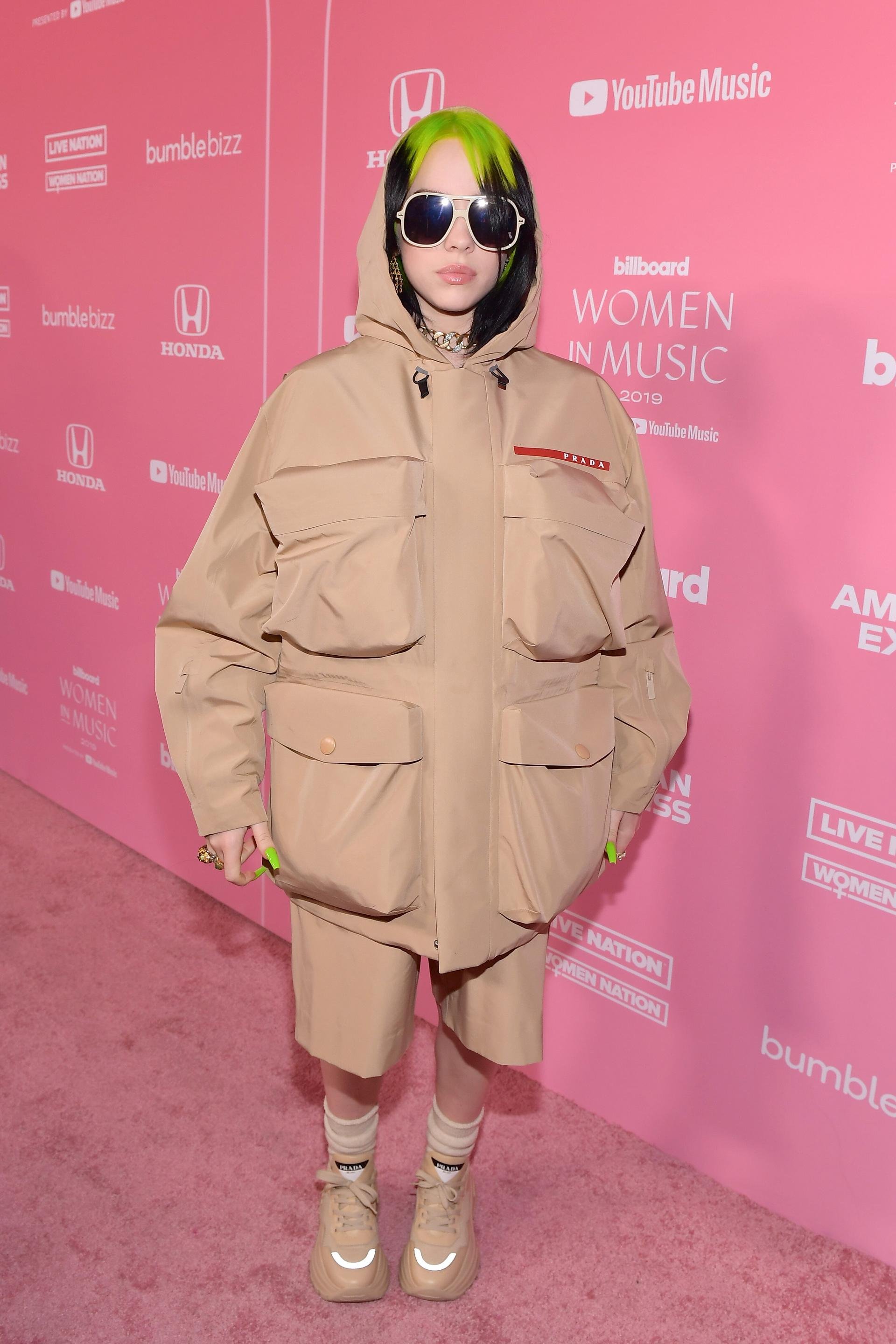 Singer Billie Eilish at Billboard Women in Music event wearing oversized tan jacket and shorts with sunglasses and green hair. 