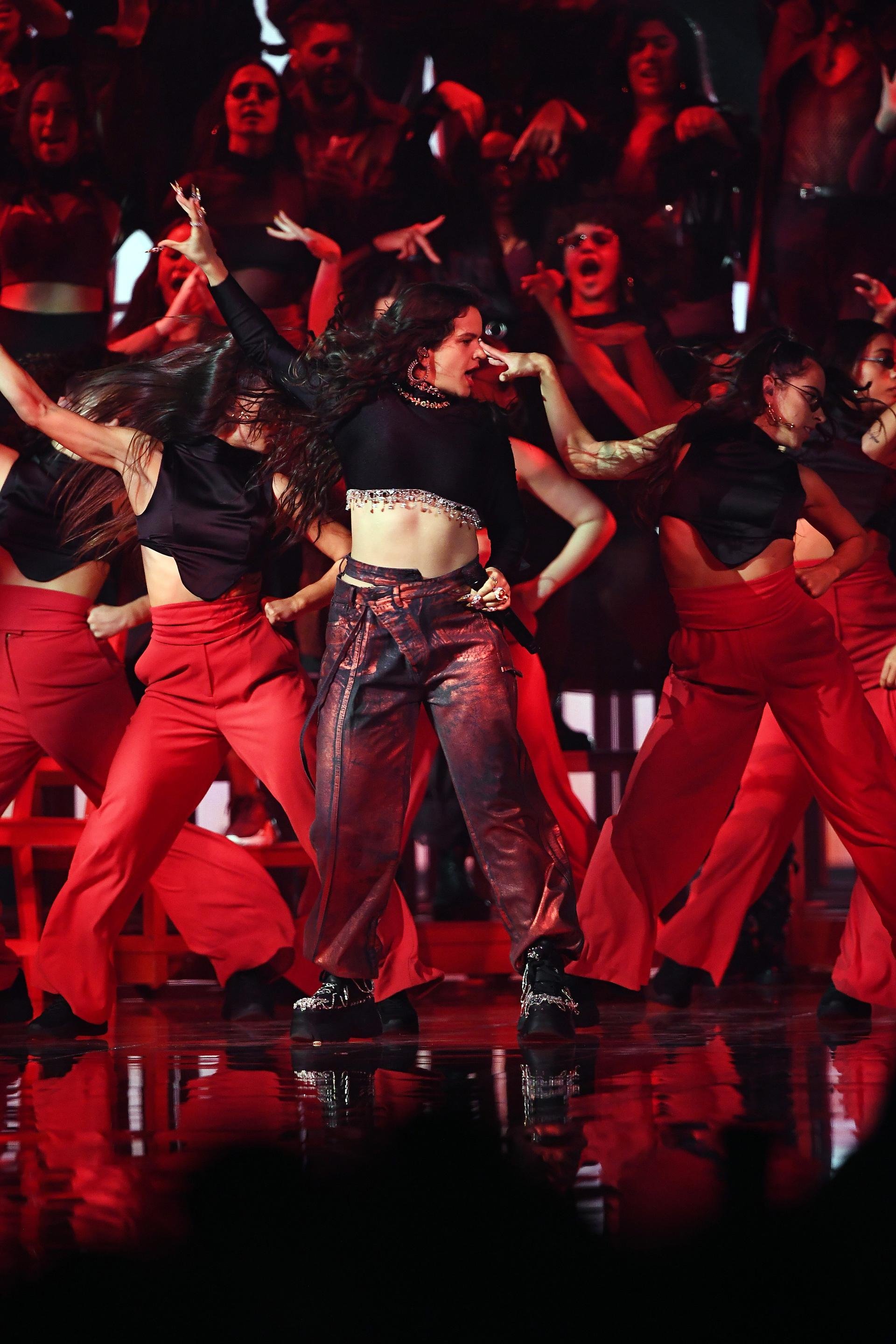 Dynamic stage performance featuring a singer surrounded by dancers in red, with dramatic lighting and reflections enhancing the energetic scene.