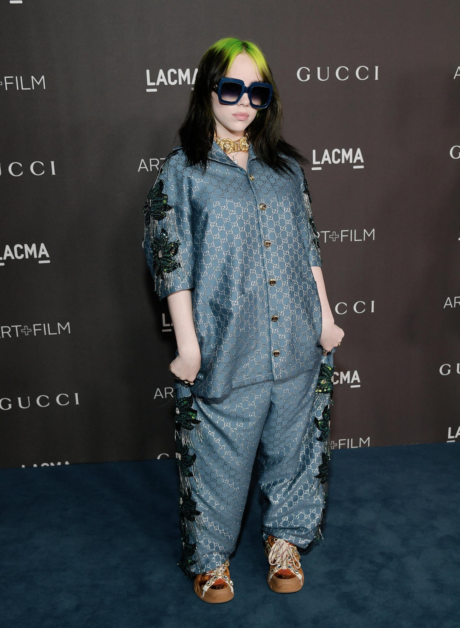 Billie Eilish stands on a dark background in an ornate, patterned, loose-fitting, turquoise Gucci suit with yellow sneakers and dark glasses.