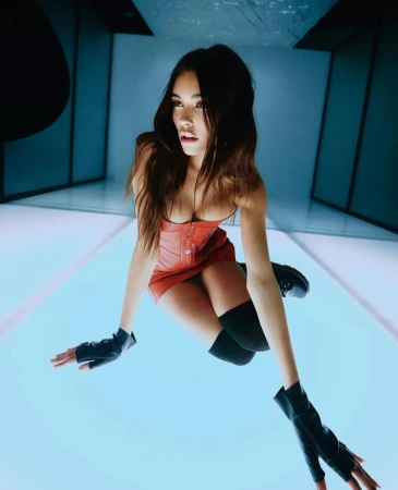 Madison Beer poses in a red corset dress, black gloves, and platform boots in a photo studio with a blue geometric backdrop.