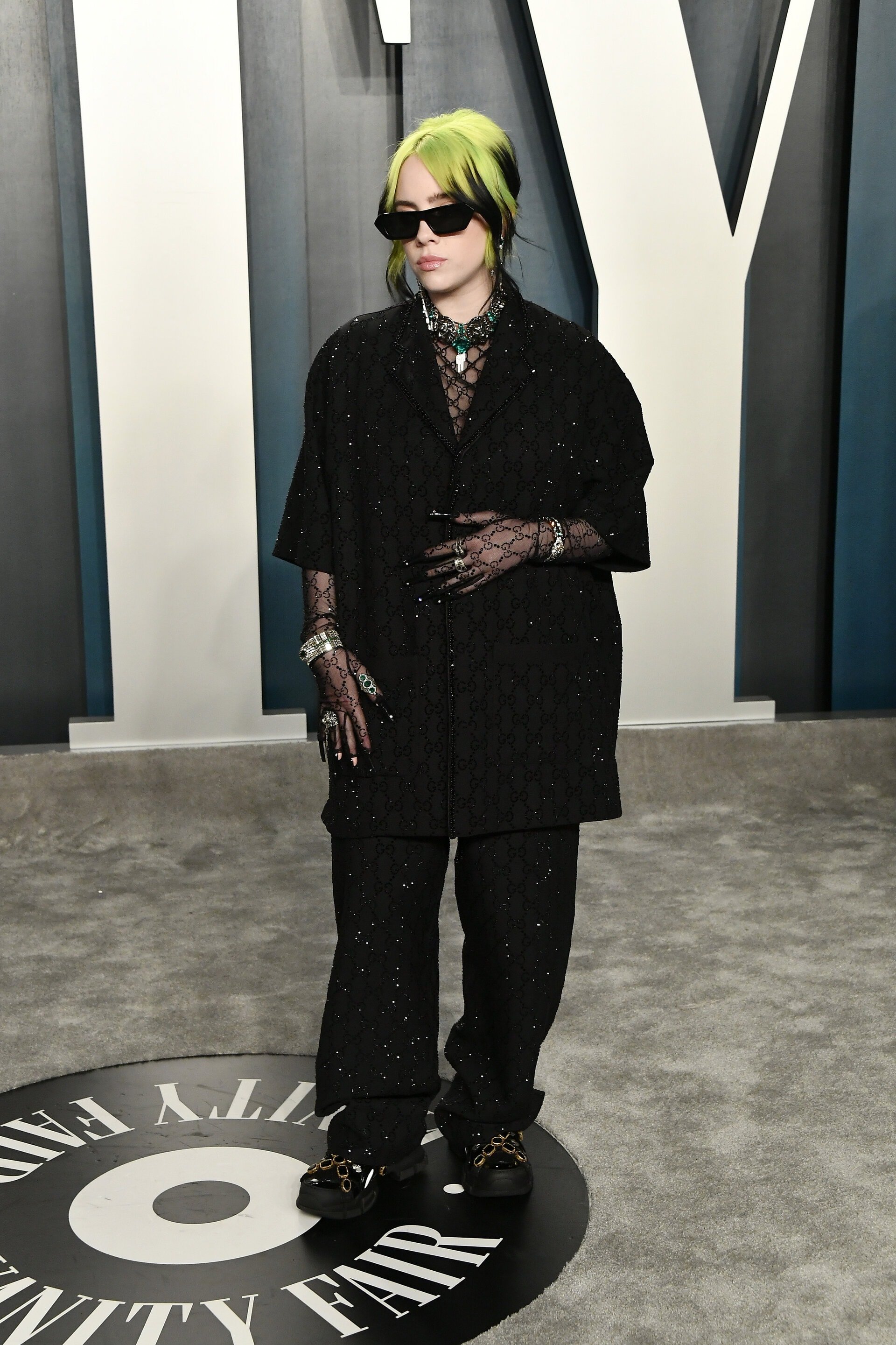 Billie Eilish poses on a Vanity Fair step-and-repeat, wearing an oversized black jacket and pants with black glasses and neon green hair.