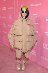 Billie Eilish wearing oversized beige hooded jacket and shorts with sunglasses standing on pink step and repeat carpet