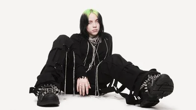 Billie Eilish sitting on a white floor wearing black clothes with green hair highlights and chunky spiked platform shoes