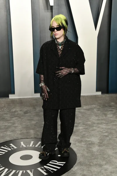 Billie Eilish standing on a branded circular floor graphic wearing an oversized black patterned suit, sunglasses, and sheer gloves.