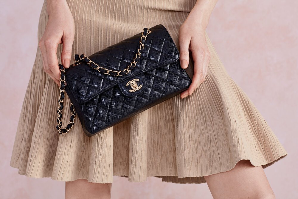 Person holds a quilted black Chanel handbag with gold chain, worn with a beige dress, set against a soft pink background.