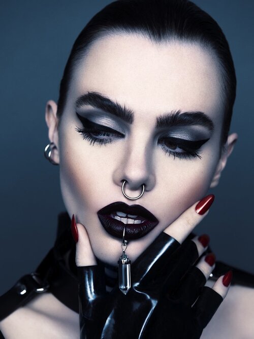 Close-up studio portrait of a woman with silver eyeshadow, dark lipstick, and multiple piercings, wearing a black latex top and gloves.