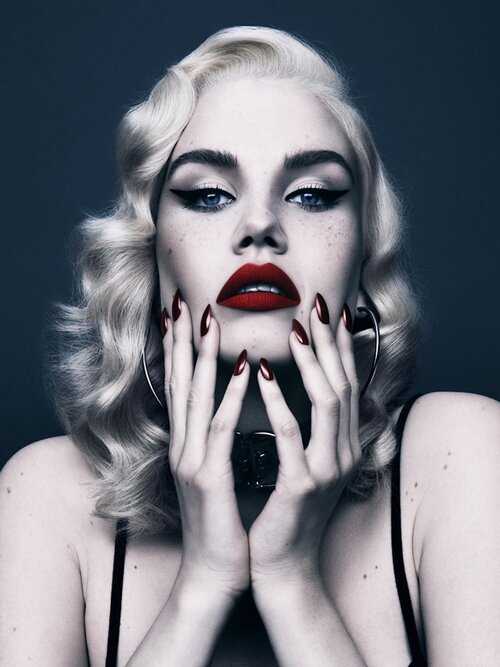 Close-up portrait of a woman with platinum blonde wavy hair, red lips, and dark eyeliner, with her hands near her face against a dark backdrop.