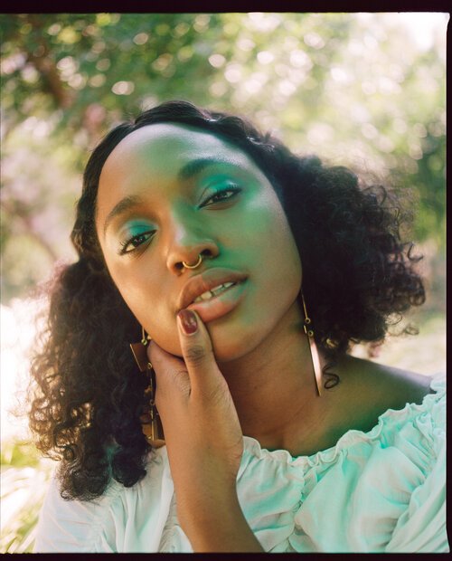 Portrait of a young Black woman with curly hair and colorful teal eye makeup wearing white top in a natural outdoor setting.