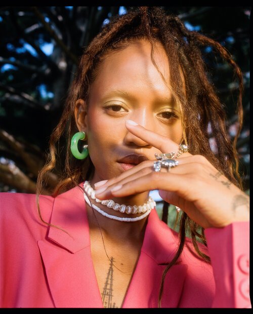 Close-up portrait of a fashionable woman with dreadlocks in a pink blazer, featuring unique makeup, jewelry, and a stylish pose outdoors.