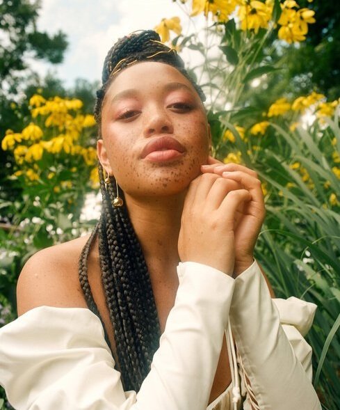 Close-up of a young Black woman posing outdoors with yellow flowers, wearing a white top and gold earrings, with braids and soft makeup.