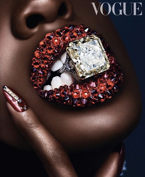 Close-up fashion beauty shot featuring a dark-skinned woman's face with a large diamond ring held in her embellished lips, Vogue cover.