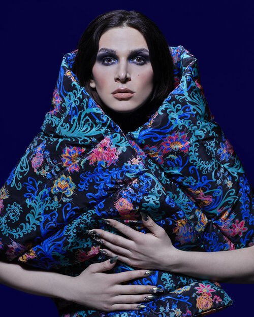 A person wearing a patterned puffer coat poses against a navy background. The coat features floral and swirling designs in blue, pink, and green.