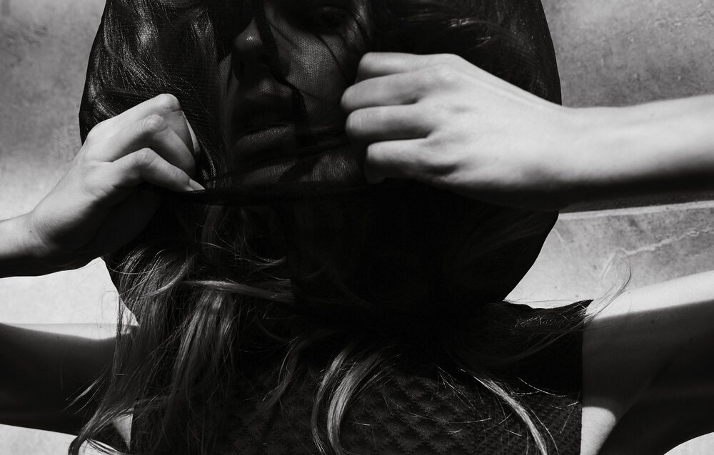 Monochrome portrait of a woman's face partially obscured by a dark veil, with her hands holding the fabric near her face, indoor setting.