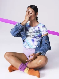 Woman sitting cross-legged wearing a tie-dye t-shirt, denim jacket, and colorful socks against a white seamless backdrop with a pink prop