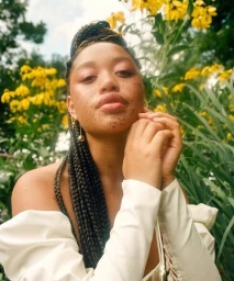 Close-up portrait of a woman with braided hair and freckles posing outdoors amidst tall green grass and yellow wildflowers.