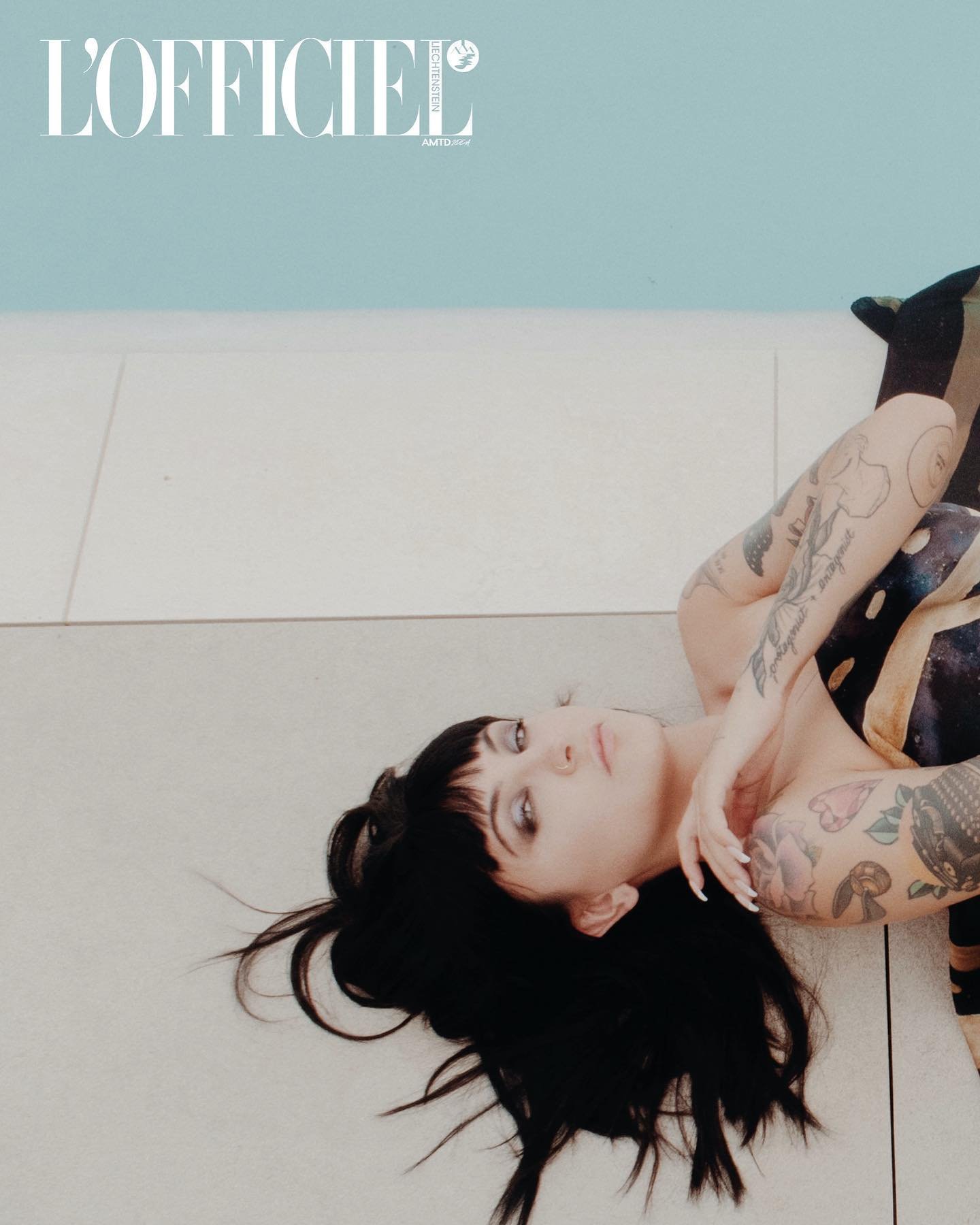 Brunette woman with tattoos lies on a white tiled floor in a close-up shot, wearing patterned leggings and posing for a fashion editorial.