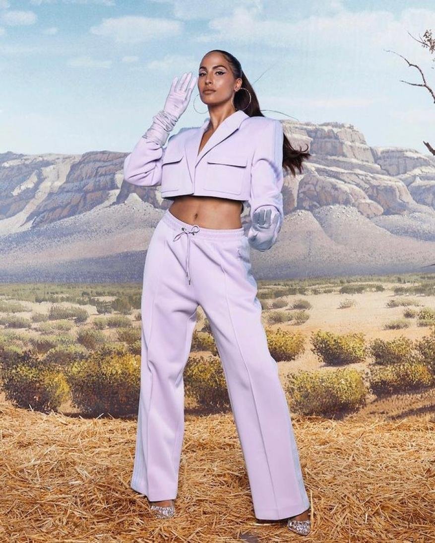 A woman in a lavender cropped suit and gloves poses against a background of mountains and plains, showcasing high-fashion apparel.