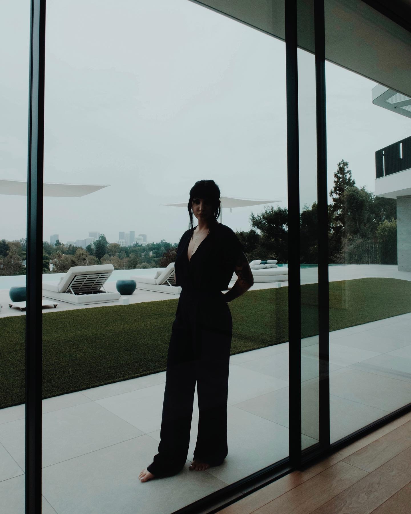 Woman in black jumpsuit stands on a patio overlooking a modern lawn with a swimming pool, lounge chairs, and city skyline.