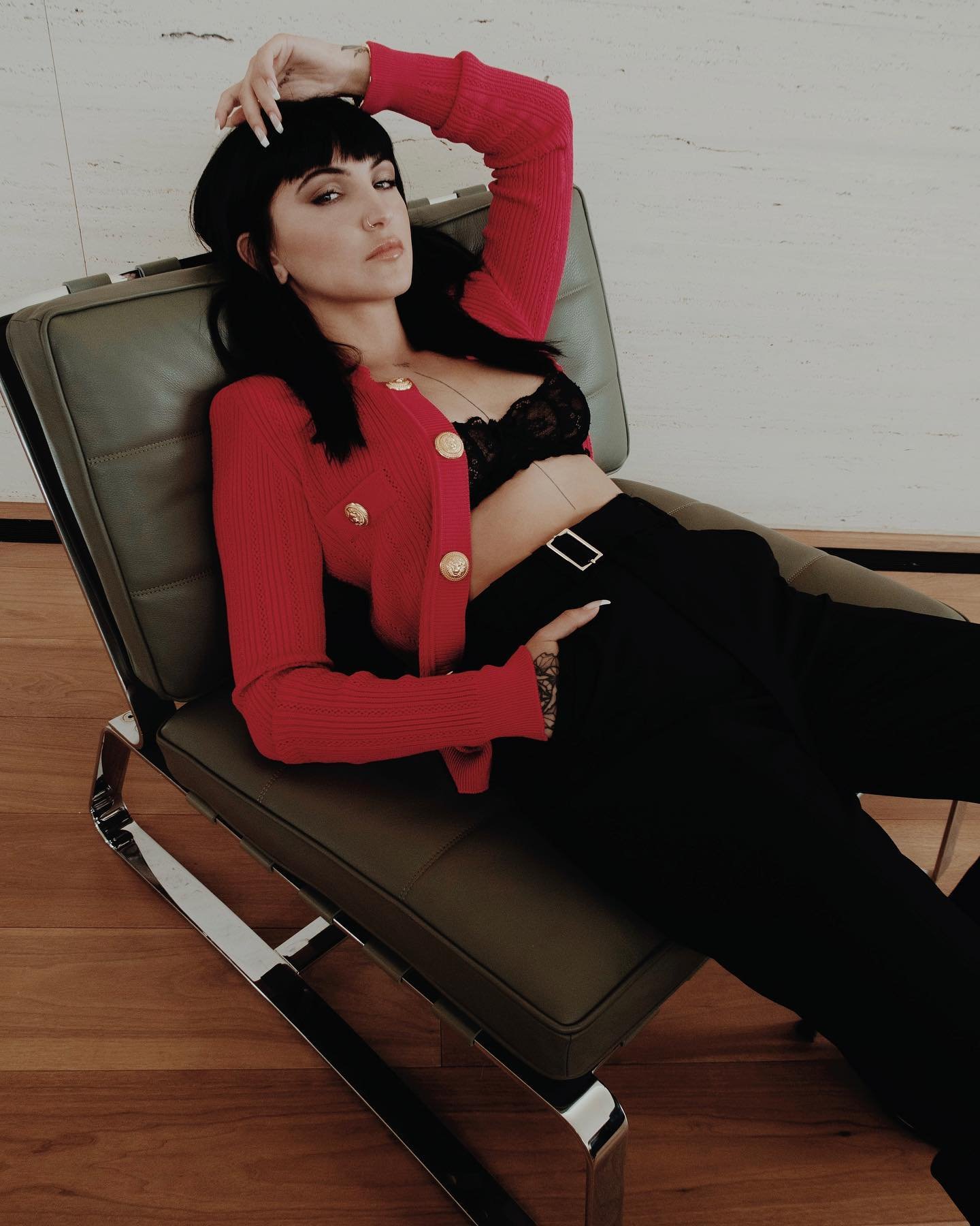 Attractive brunette in a red cardigan and black lingerie and pants poses in a chair on a wood floor in a brightly lit space.