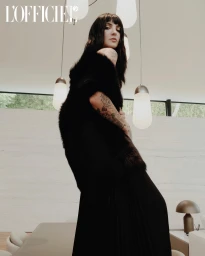 A woman with dark hair and bangs wearing a black fur stole and long pleated skirt in an interior space