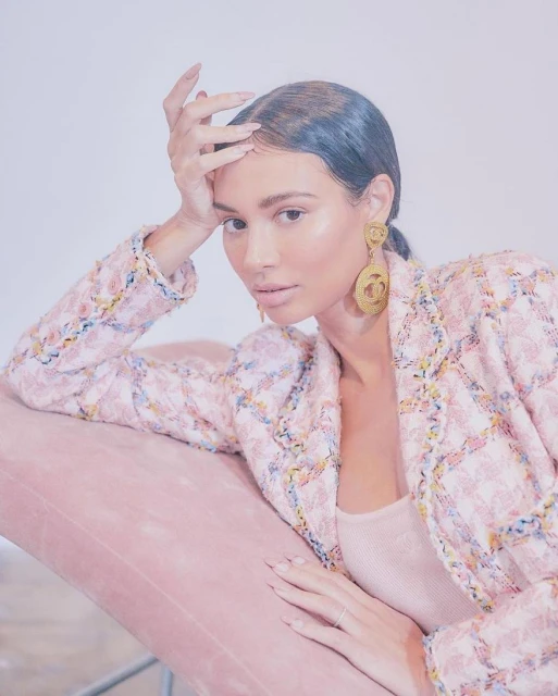 Portrait of a woman with shoulder-length hair, posing on a pink chair in a pastel jacket and large gold earrings in a bright indoor setting.