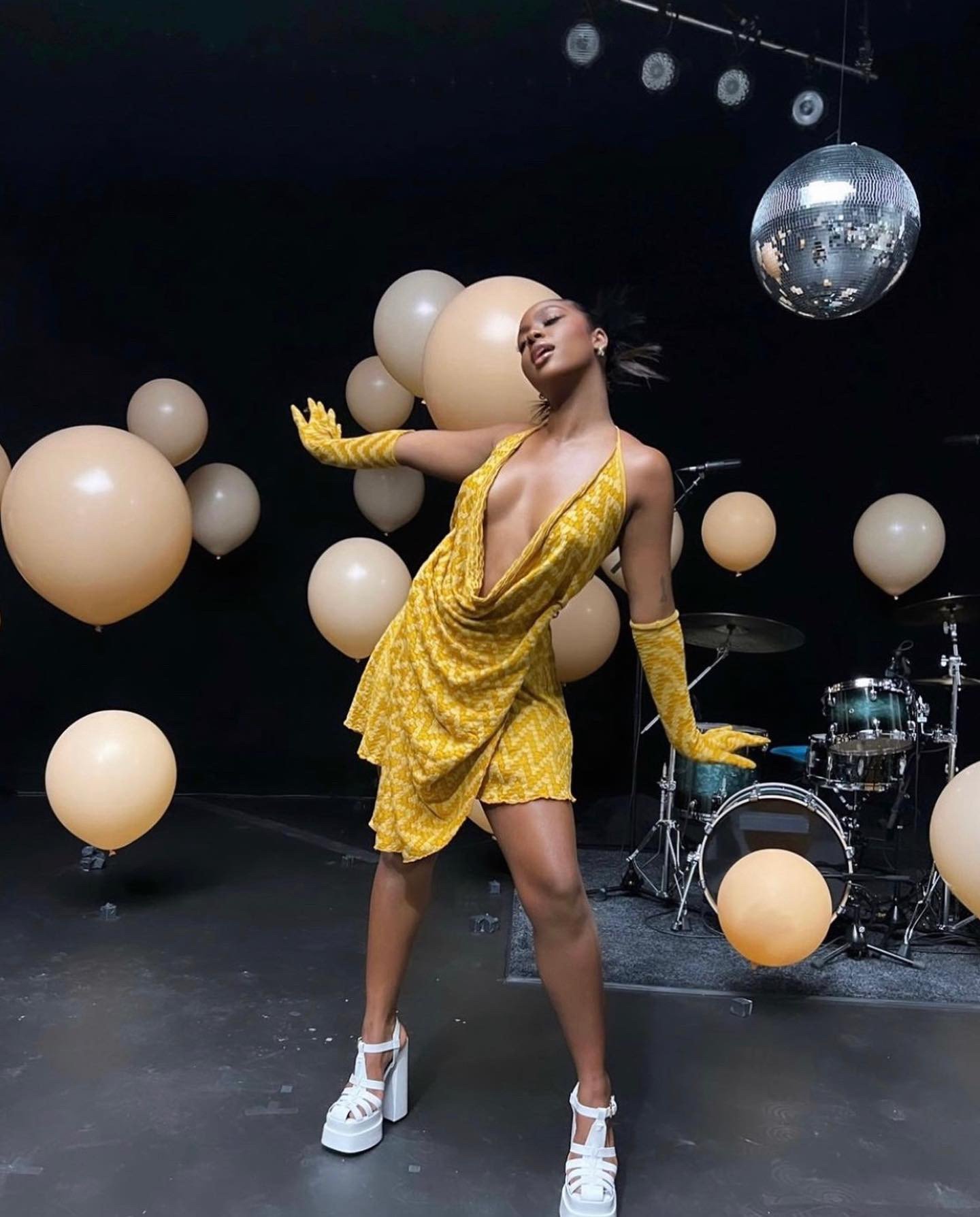 A woman in a yellow dress with balloons and a disco ball stands in front of a drum set in a dark room. The balloons add a playful touch.