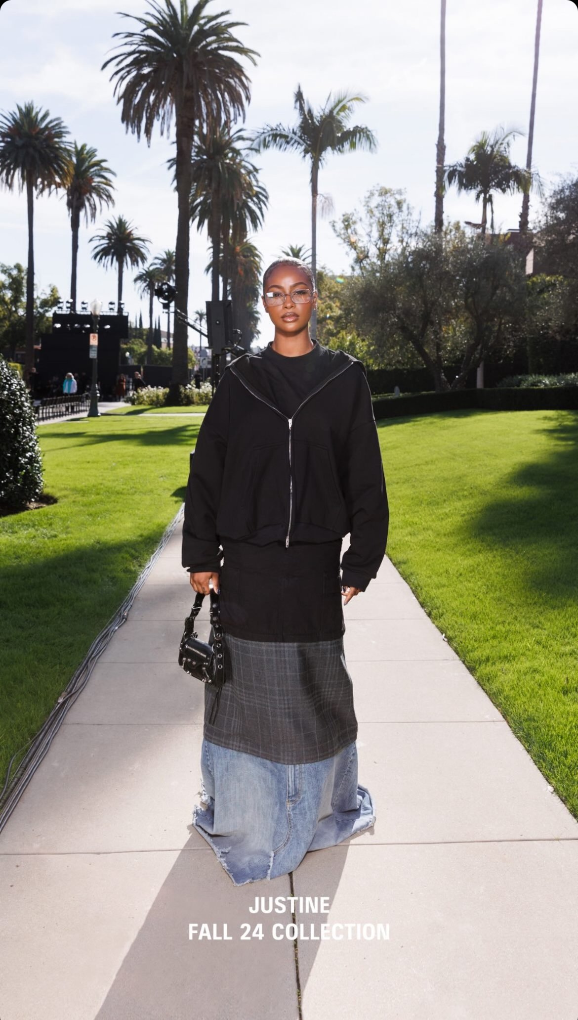 Woman in a black jacket and multi-layered denim skirt walks on a path lined with palm trees on a sunny day.