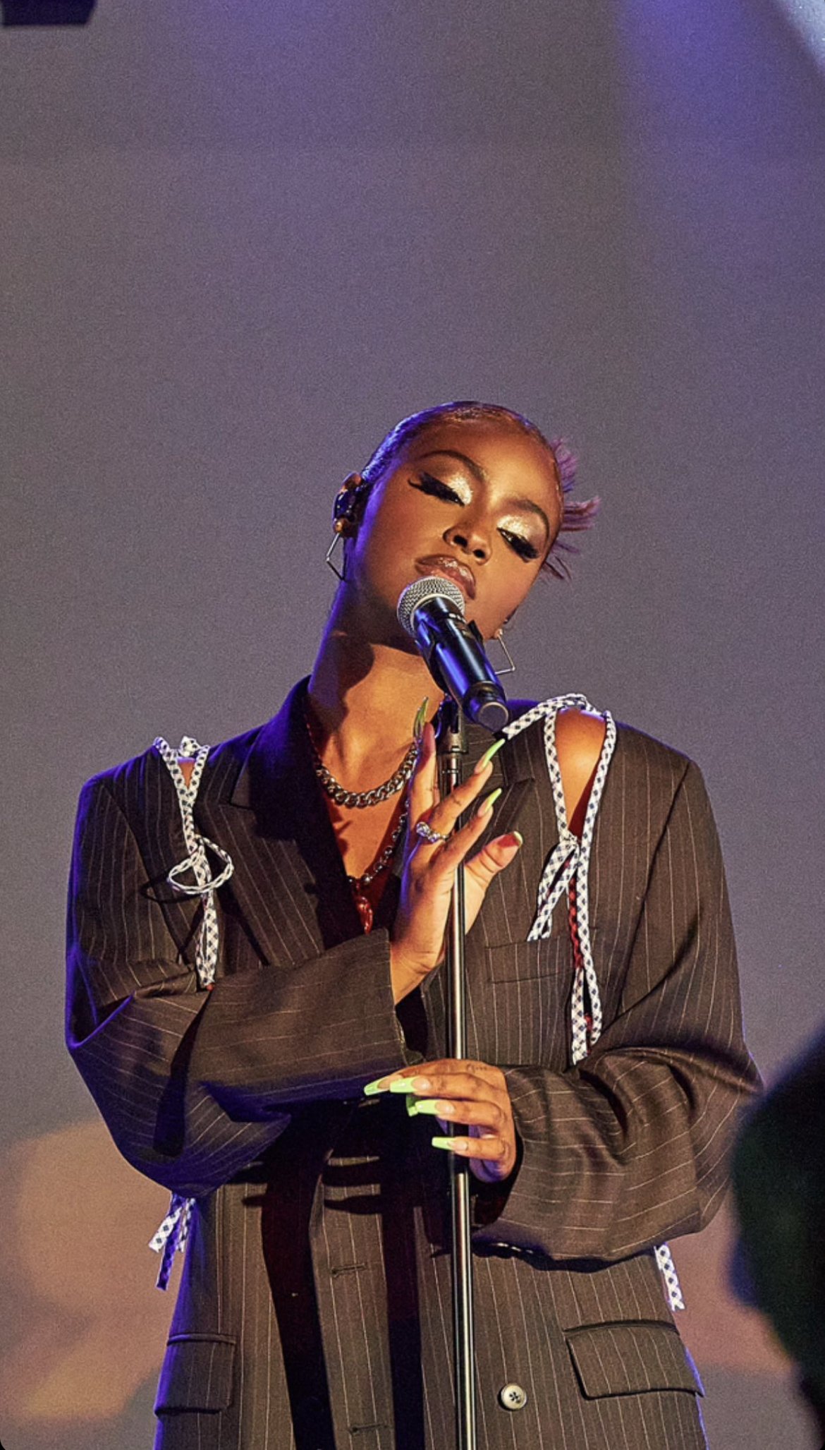 African-American woman sings on stage wearing a pinstripe blazer with open shoulders, jewellery, and green nails, with a microphone.