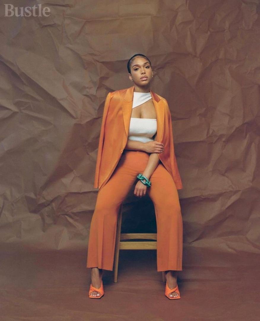 A stylish woman with orange blazer and pants sits on a chair against a textured brown backdrop in a fashion editorial shot.