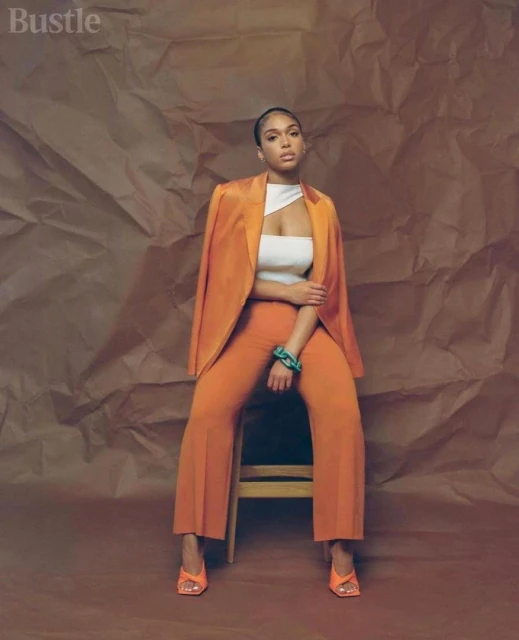 A stylish woman with orange blazer and pants sits on a chair against a textured brown backdrop in a fashion editorial shot.