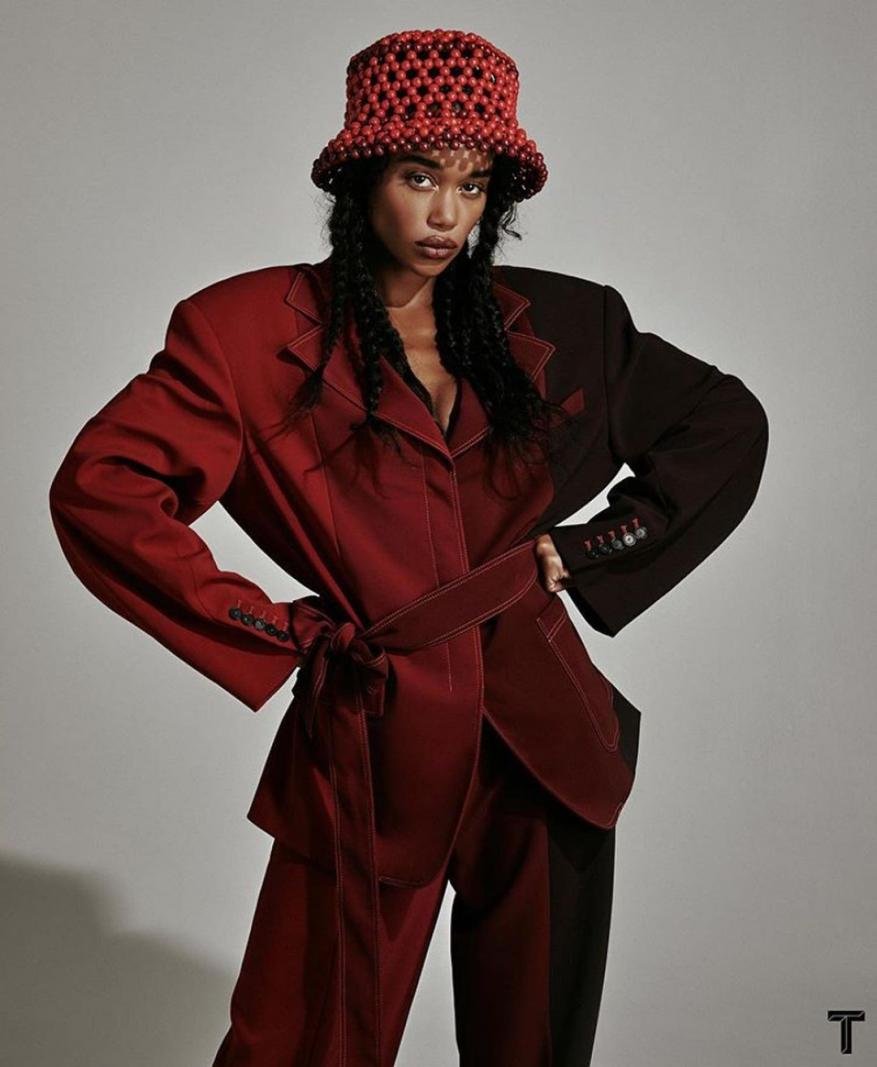 A young woman poses in a two-tone jacket and bucket hat, hands on hips, against a grey studio background. Fashion portrait. 