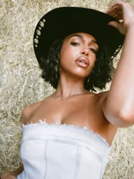 Close-up portrait of a woman wearing a black embellished cowboy hat and a light denim strapless top against a hay bale background.