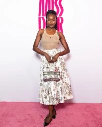 Black woman wearing sheer beige top and patterned skirt holding a branded handbag in front of a pink backdrop