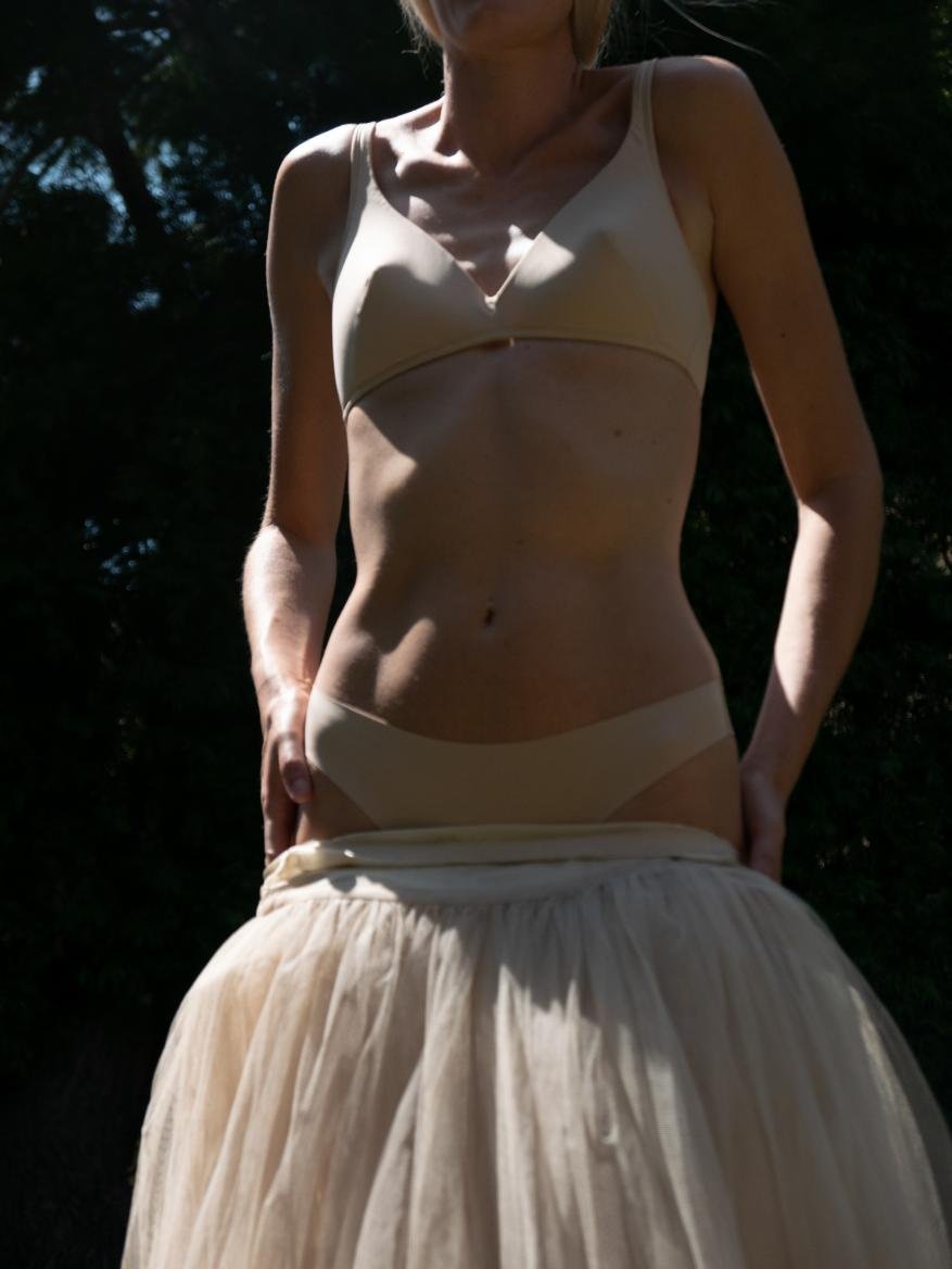 Woman wearing beige bra and underwear, with an oversized tulle skirt around her waist, standing outdoors against a dark green backdrop.