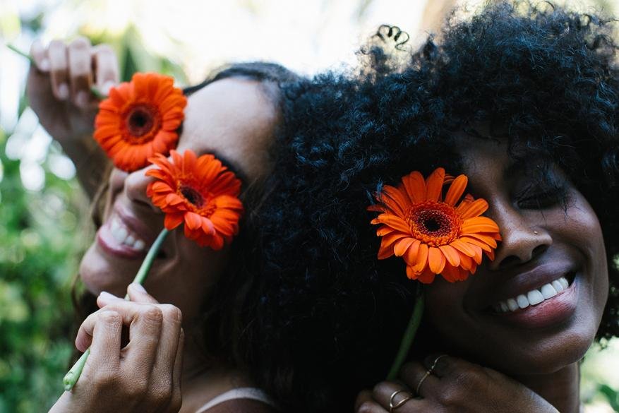 Two smiling women pose outdoors with bright orange gerbera daisies covering their eyes in a close-up shot, showing youthful joy.