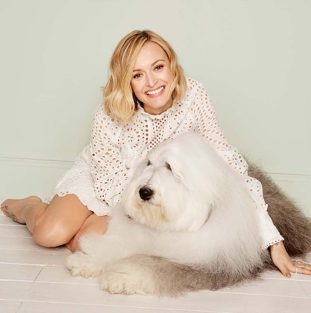 Smiling woman in eyelet dress sits on the floor with a large Old English Sheepdog in a bright indoor setting, creating a cozy, inviting scene.