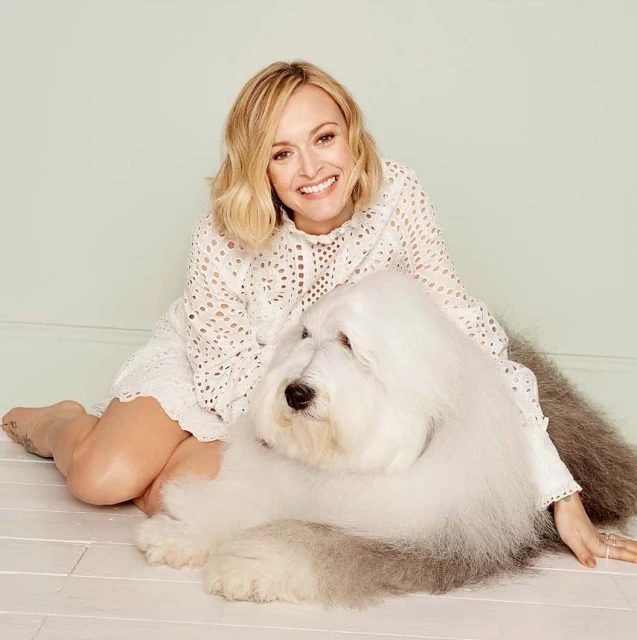 Smiling woman in eyelet dress sits on the floor with a large Old English Sheepdog in a bright indoor setting, creating a cozy, inviting scene.