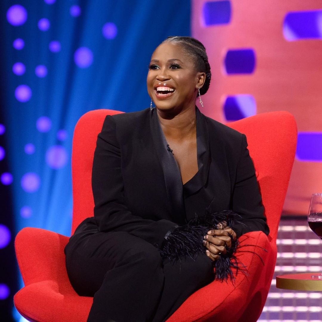 Motsi Mabuse, smiling and sitting on red chair in a black pantsuit on TV show set with a bright blue and purple backdrop.