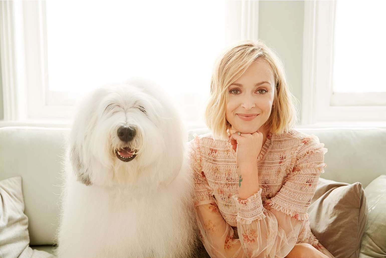 Blonde woman wearing a patterned pink top sitting on a sofa next to a large fluffy Old English Sheepdog.