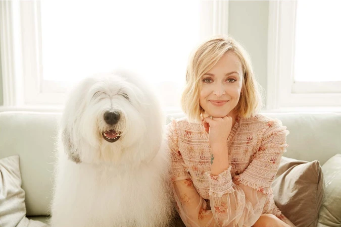 Blonde woman wearing a patterned pink top sitting on a sofa next to a large fluffy Old English Sheepdog.