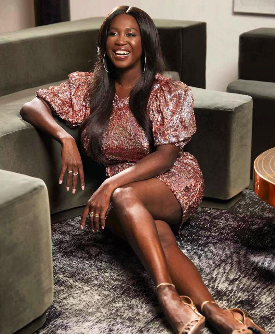 Smiling Black woman in a sequined dress seated indoors. She is wearing strappy heels, and the background includes a sofa and patterned rug.