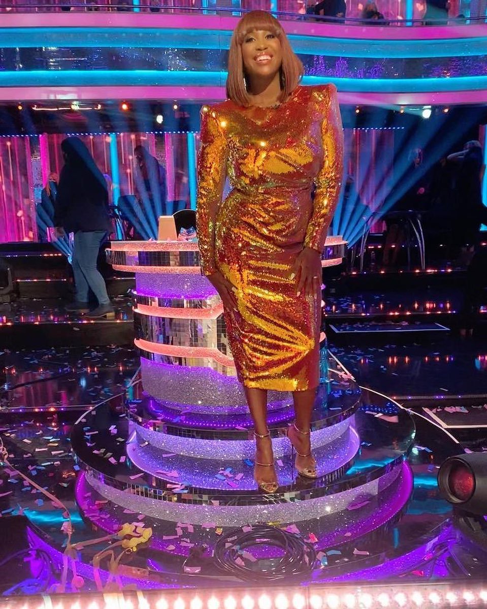 Woman in a gold sequin dress standing on a stage with confetti and lights, smiling and looking forward, performing at a music event.