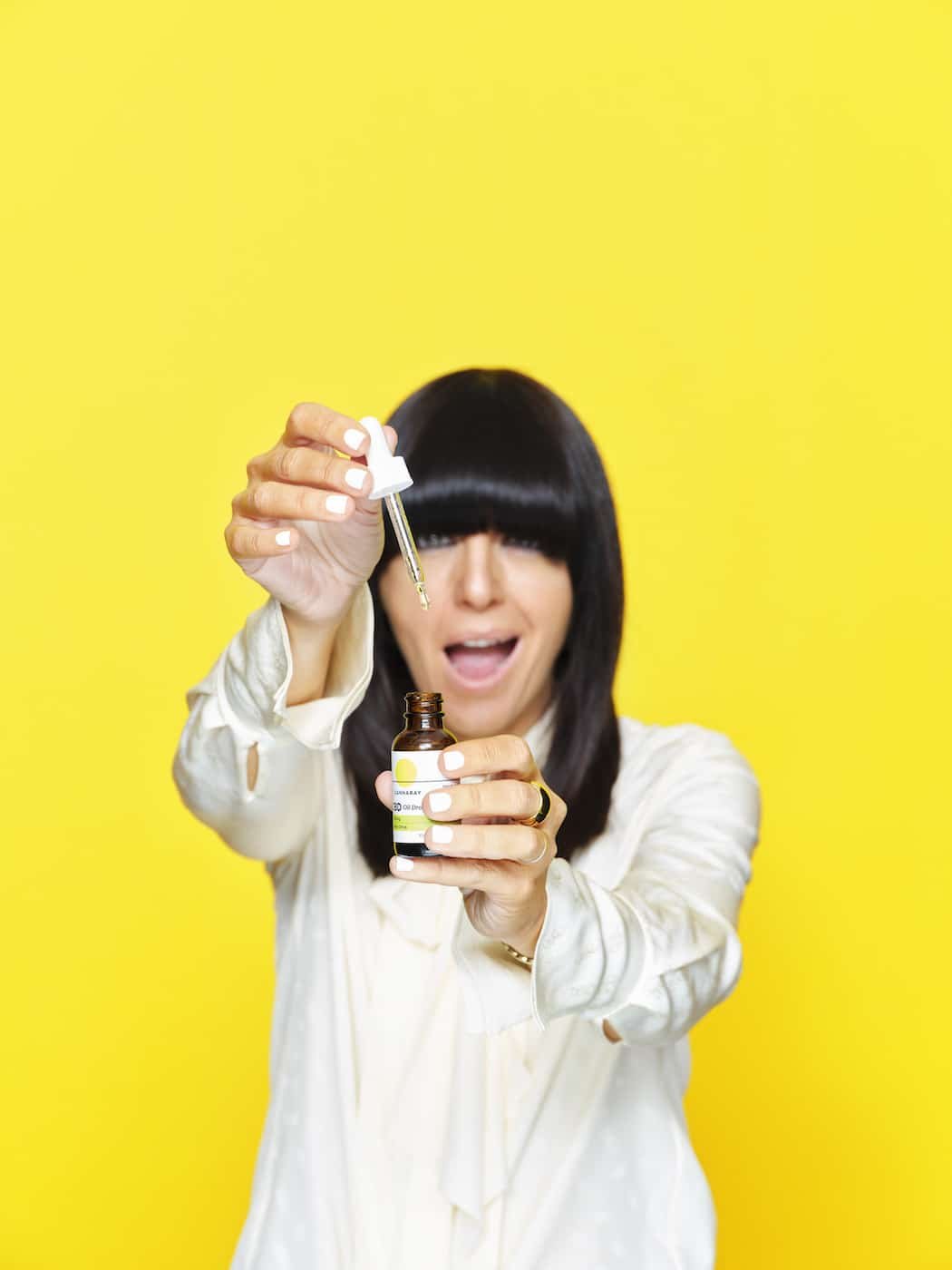 Woman with dark hair smiles, holding cosmetic serum bottle with dropper. Bright yellow background, focus on serum and joyful expression.