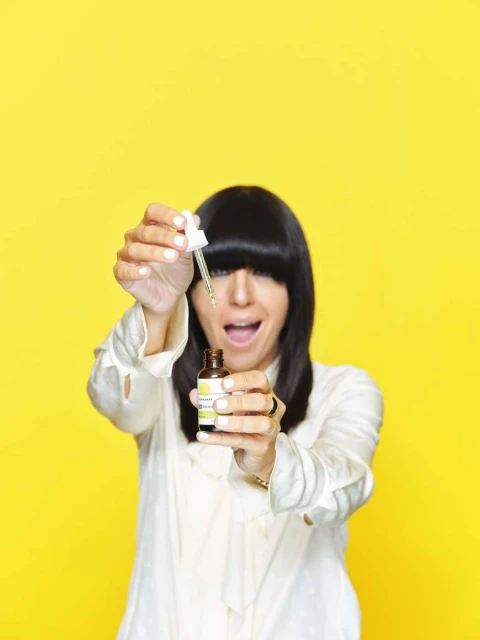 Woman with dark hair smiles, holding cosmetic serum bottle with dropper. Bright yellow background, focus on serum and joyful expression.