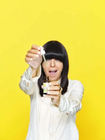 Woman with dark hair smiles, holding cosmetic serum bottle with dropper. Bright yellow background, focus on serum and joyful expression.